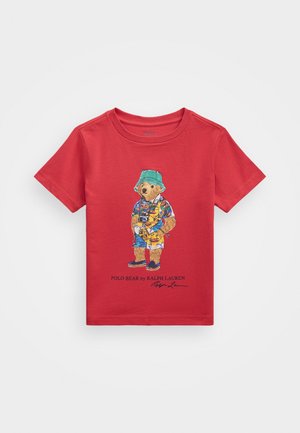 Punane puuvillane T-särk, millel on värvikas karu graafika, kes kannab rannariideid, kaamerat ja sandaale, ning tekst "Polo Bear by Ralph Lauren".