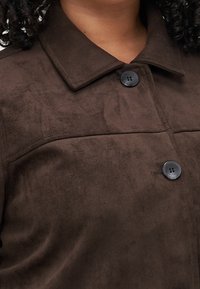 Hnědá bunda z umělé semiše s límečkem, dvěma černými knoflíky a hladkým, texturovaným povrchem.