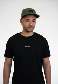 Fekete póló "JOHNNY URBAN" felirattal fehér színben, zöld és fekete sapkával, fekete folttal, sima fehér háttér előtt.
