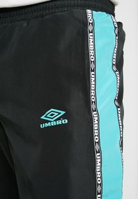 Sorte træningsbukser med et sidepanel i lyseblå og et sort-hvidt logo-stribe. Materialet ser glat ud med et elastisk waistband.