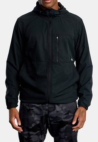 Sort hættetrøje lavet af letvægtsmateriale, med en lynlåslomme på brystet, elastiske manchetter og et enkelt design, parret med camouflagebukser.