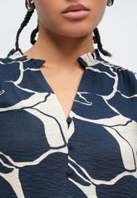 Bluza bleumarin cu un model abstract crem, decolteu în V, guler cu volane și șase nasturi acoperiți cu material pe partea din față.