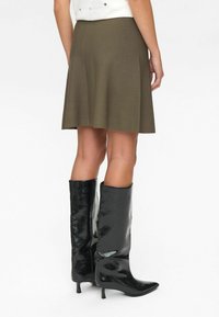 Femme portant une jupe olive longueur genoux et des bottes noires à bout pointu montantes avec de petits talons, se tenant contre un fond blanc.