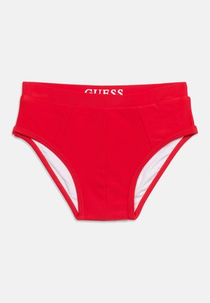 Bas de bikini taille haute rouge avec doublure intérieure blanche et logo "GUESS" visible sur la ceinture intérieure sur fond uni.