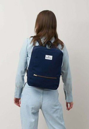 Mochila de lona azul marino con un diseño plano, detalles en cremallera dorada y un bolsillo frontal. Parche del logo en la parte superior central de la mochila.