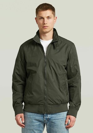 Jonge man die een donkergroen jack met ritssluiting draagt, met geribbelde manchetten en tailleband, staand tegen een effen lichte achtergrond.