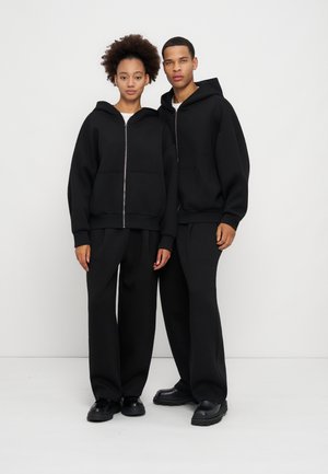 Un hombre y una mujer están de pie uno al lado del otro, vestidos con sudaderas negras con cremallera a juego, pantalones negros holgados y zapatos negros sobre un fondo blanco.