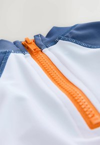 Hvid og blå zip-up beklædning med en orange lynlås. Blødt, glat stof med detaljeret syning langs kanterne.