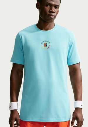 Uomo che indossa una t-shirt Nike Court azzurra con logo multicolore, pantaloncini arancioni e fasce Nike bianche su entrambi i polsi.