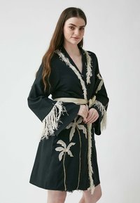 Svart kappa med intrikata vita palmlöv broderier och fransdetaljer på ärmarna. Höjdpunkter inkluderar en v-ringning och ett knytbart midjeband.