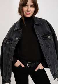 Czarna kurtka dżinsowa z zapięciem na guziki, dwoma kieszeniami na piersi i luźnym krojem. Noszona na czarnym golfie, w połączeniu z czarnymi spodniami i paskiem.