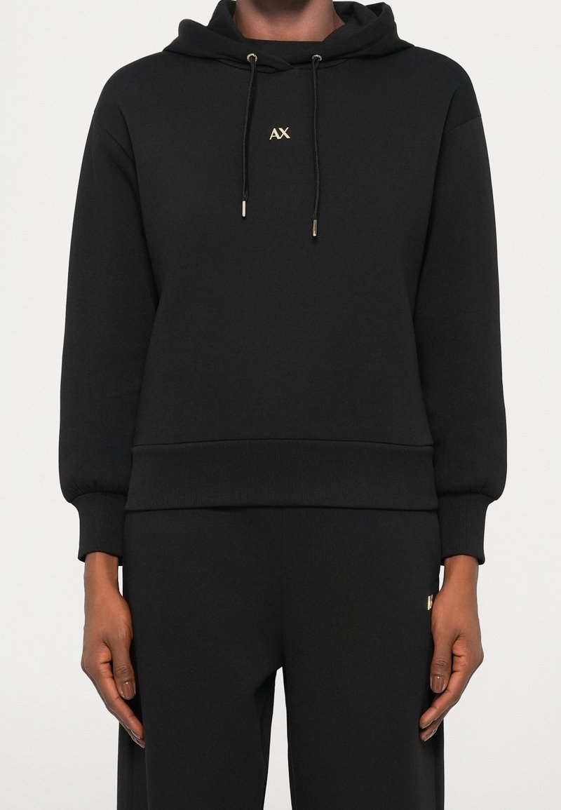 Sweat à capuche noir avec logo doré "AX" sur la poitrine, cordons avec embouts métalliques, porté par une personne les mains détendues sur les côtés.