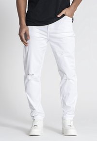 Vaqueros blancos desgastados con roturas en las rodillas, hechos de tela vaquera. Combinados con zapatillas blancas y una camiseta negra. Diseño sencillo.