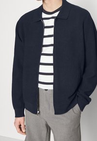 Námořnická žebrovaná bunda na zip s límečkem, která má texturovaný design, nošená přes pruhované tričko a šedé kalhoty.