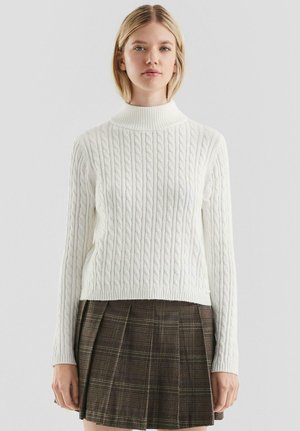 Jonge vrouw die een witte kabelgebreide coltrui en een bruin geruite geplooide rok draagt, staande tegen een effen achtergrond.