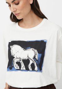 Mujer vistiendo una camiseta blanca con un boceto abstracto en negro y azul de un caballo en la parte frontal, con la cabeza ligeramente inclinada hacia abajo.