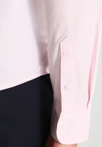 Jasnoróżowy mankiet długiego rękawa z zapięciem na guzik na ciemnym tle.