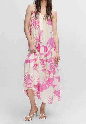 Kobieta ubrana w bezrękawnikową, sięgającą kostek kremową sukienkę w różowe tropikalne liście oraz białe sandały na obcasie, trzymająca część sukienki.