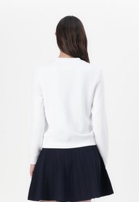 Felpa bianca con maniche lunghe, scollo rotondo e polsini a coste indossata sopra una gonna plissettata blu navy. Il tessuto appare liscio e informale.