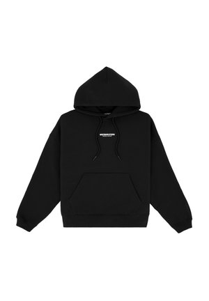 Jeune homme portant un sweat à capuche noir avec un petit texte blanc, un jean bleu clair ample, des chaussures noires et de grandes lunettes de soleil noires, debout sur un fond gris.