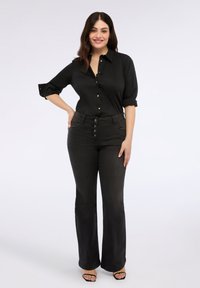 Camicia nera con bottoni, con maniche arrotolate, abbinata a jeans svasati alti in vita di colore grigio scuro, con dettagli dei bottoni e una texture liscia.