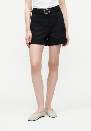 Naine, kes kannab musti mansettidega lühikesi pükse musta vööga, valget topi ja beeže madalate kontsadega jalanõusid, seisab tavalise valge tausta ees.