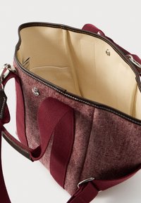 Burgundi színű, puha textúrájú anyagú kézitáska, világos belső térrel, több rekesszel és barna bőr díszítésekkel.