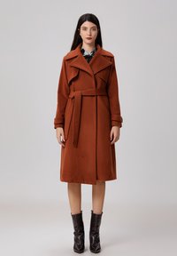 Long manteau en laine marron au design trench, avec une taille ceinturée, de larges revers et une texture douce, porté sur une chemise à motifs et associé à des bottes brillantes.