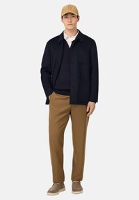 Vyras, vilkintis šviesios rudos spalvos kepurę, tamsiai mėlyną striukę ant tamsaus megztinio ir baltos marškinių, rudos kelnės ir taupe zomšos batus, stovi su viena ranka kišenėje.