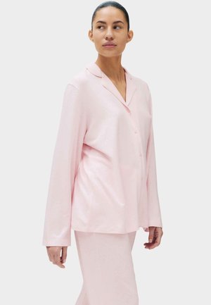 Donna con pigiama rosa pallido con bottoni e pantaloni abbinati, in piedi e che guarda leggermente verso destra.