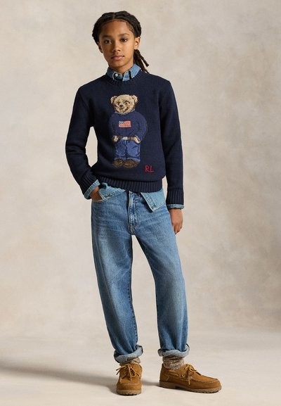 Jeune personne aux cheveux tressés porte un pull bleu marine avec un motif d'ourson et de drapeau, une chemise en jean, un jean large, manches retroussées, chaussures marron et chaussettes.