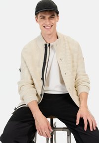Beige strikket cardigan med ribbet struktur, med en sort lynlås og knapper, båret over en hvid skjorte og sorte bukser.