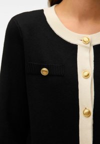 Cardigan nero in maglia con rifiniture e colletto color crema, dotato di bottoni tondi dorati e una decorativa patta nera della tasca con un bottone dorato.