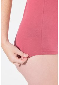 Abbigliamento in cotone rosa con un primo piano sull'orlo e la cucitura laterale. Una mano afferra il tessuto, mostrando la sua morbida texture e l'elasticità.