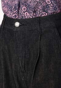 Black denim trousers with a high waist, featuring a metallic button closure, subtle texture, and front pleats, paired with a patterned top.
