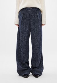 Calças de perna larga feitas de tecido azul marinho texturizado com padrões subtis, apresentando pregas frontais e um fecho de botão. Calçado preto visível.