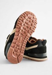 Czarne buty sportowe mają teksturowaną podeszwę z różowymi wypustkami, beżowy akcent wzdłuż boku oraz smukłą czarną cholewkę z siateczkowymi detalami.