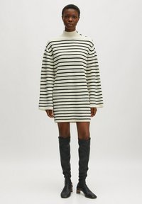 Robe tricot rayée en crème et noir, avec un col haut, des manches longues et une coupe ample, associée à des bottes noires au-dessus du genou.