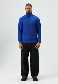 Blauwe gebreide coltrui met een gestructureerd patroon, gecombineerd met losse zwarte broek en zwarte schoenen met opvallende zooldetails.