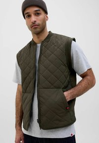 Quiltet olivengrøn vest med lynlås, rund halsudskæring og sidelommer, båret over en lys grå t-shirt og mørke jeans.