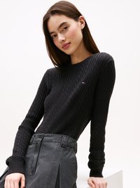 Maglione nero a coste con scollo rotondo e polsini a costine, indossato con una gonna grigia plissettata. Dettaglio di piccolo logo sul petto.