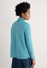 Pull en maille de couleur bleu clair avec un col rayé vert et blanc. Présente des fentes sur les côtés à l'ourlet et des manches longues.
