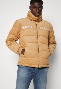 Karamellfarget cordfløyelsdunjakke med høy krage, har quiltede seksjoner og hvit "KARL"-logo på brystet. Glidelås, elastiske mansjetter.