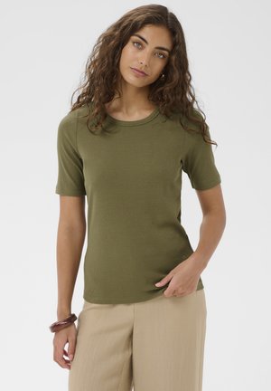 Sieviete ar cirtainiem brūniem matiem, tērpusies olīvu zaļā īsroku krekliņā un bēšās biksēs, stāv pret vienkāršu gaišu fonu.