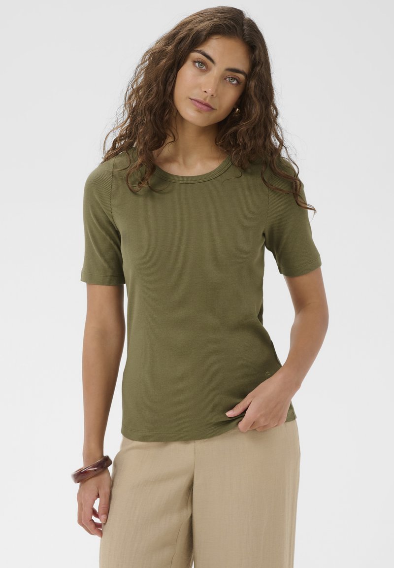 Vrouw met krullend bruin haar, gekleed in een olijfgroen shirt met korte mouwen en beige broek, staand tegen een eenvoudige lichte achtergrond.