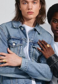 Veste en denim avec un lavage bleu clair ; modèle portant un t-shirt blanc en dessous. Gros plan sur des mains mettant en valeur des bagues en or sur les doigts.