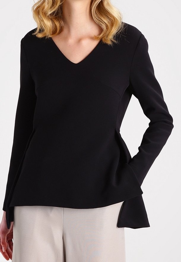 Top nero a maniche lunghe con scollatura a V, design strutturato, orlo a peplo e texture liscia. Abbinato a pantaloni di colore chiaro.