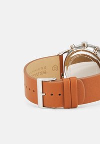 Cinturino in pelle marrone con fibbia in acciaio inox argentato. Presenta pelle texturizzata e una fodera interna liscia. Quadrante dell'orologio rotondo visibile.