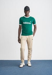 Πράσινο μπλουζάκι Adidas με λευκές λεπτομέρειες και λογότυπο, μπεζ φόρμα με μαύρες ρίγες στο πλάι και λευκά παπούτσια Adidas, συνδυασμένα με μαύρο καπέλο.