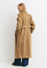 Un long manteau beige fabriqué en matériau doux, avec une taille ceinturée, de grandes poches et un col classique.